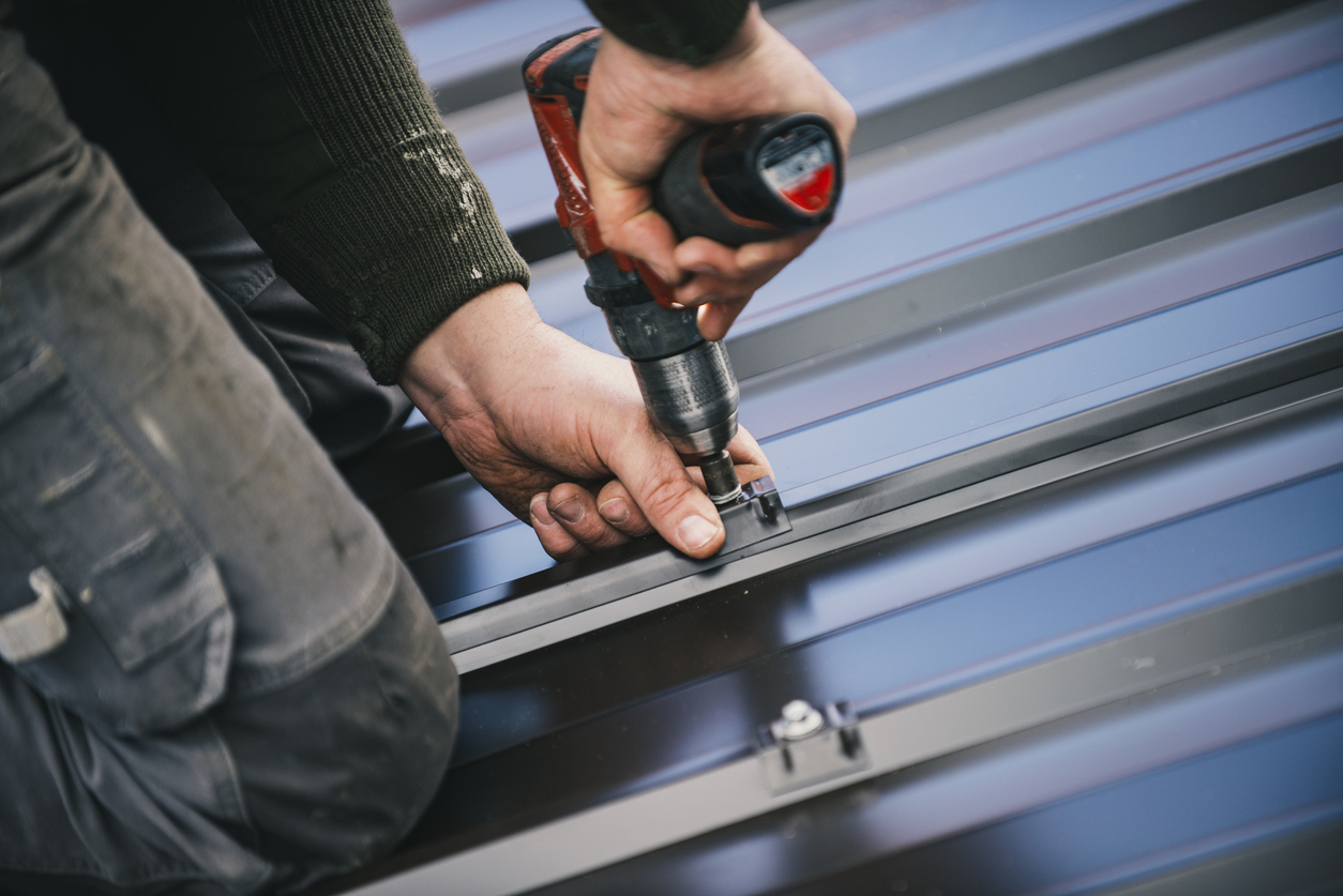 PD Home Exterior crew member is installing metal roofing sheets on the rooftop of the house using electric screwdriver.